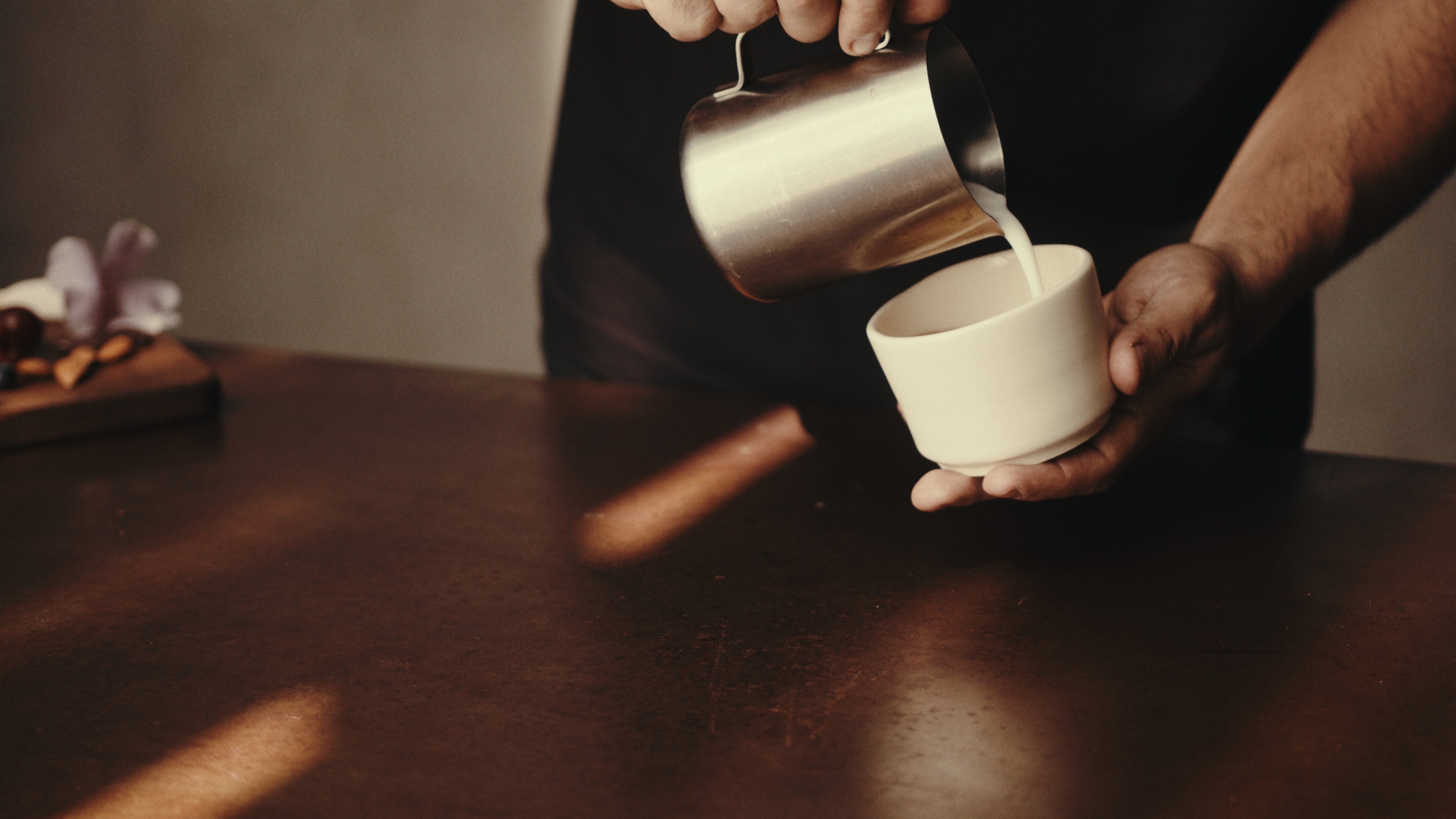 Person pouring milk from a silver pitcher into a KAFE coffee cup on a wooden table.