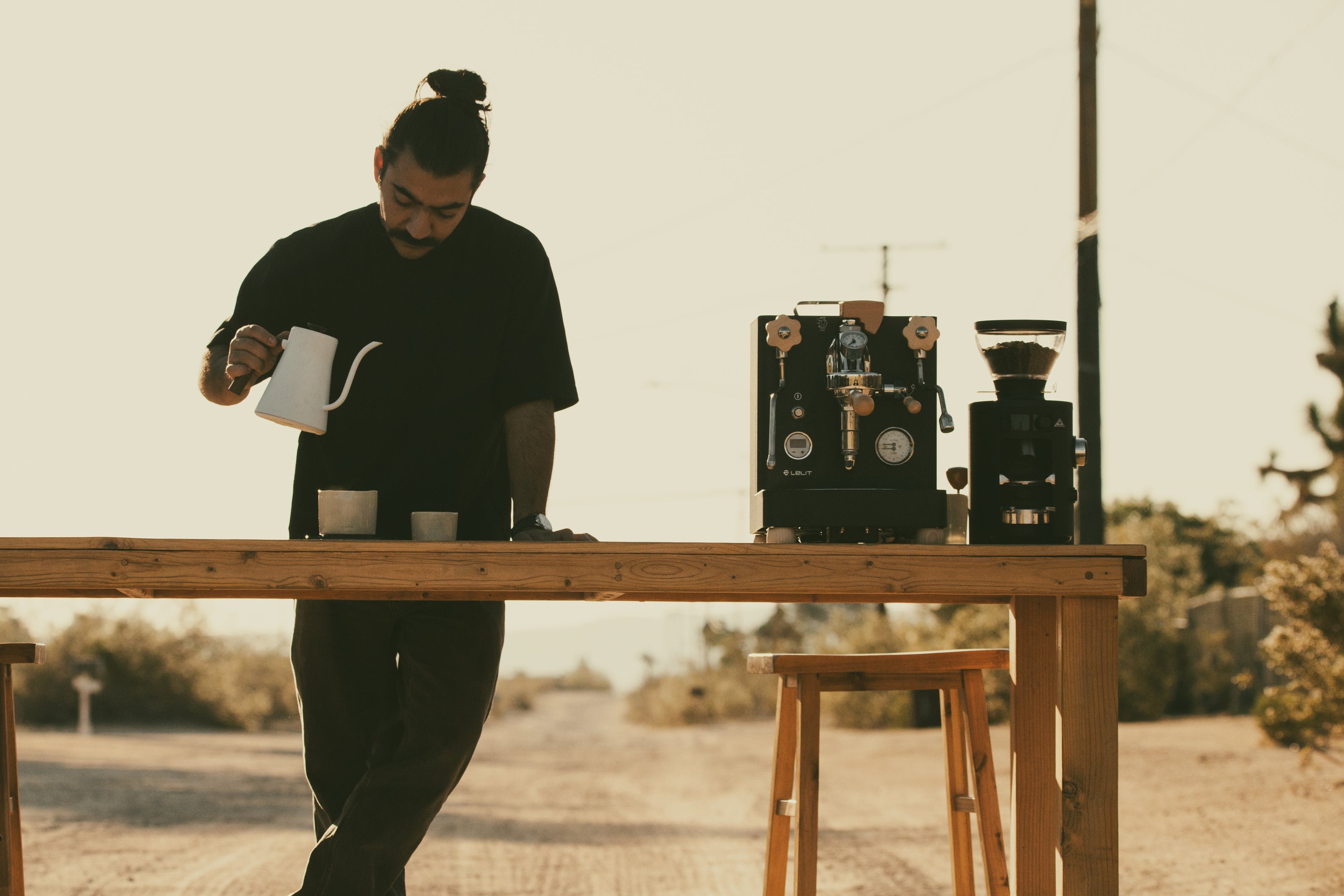 Person preparing KAFE coffee at a wooden table outdoors with a coffee machine in the background.