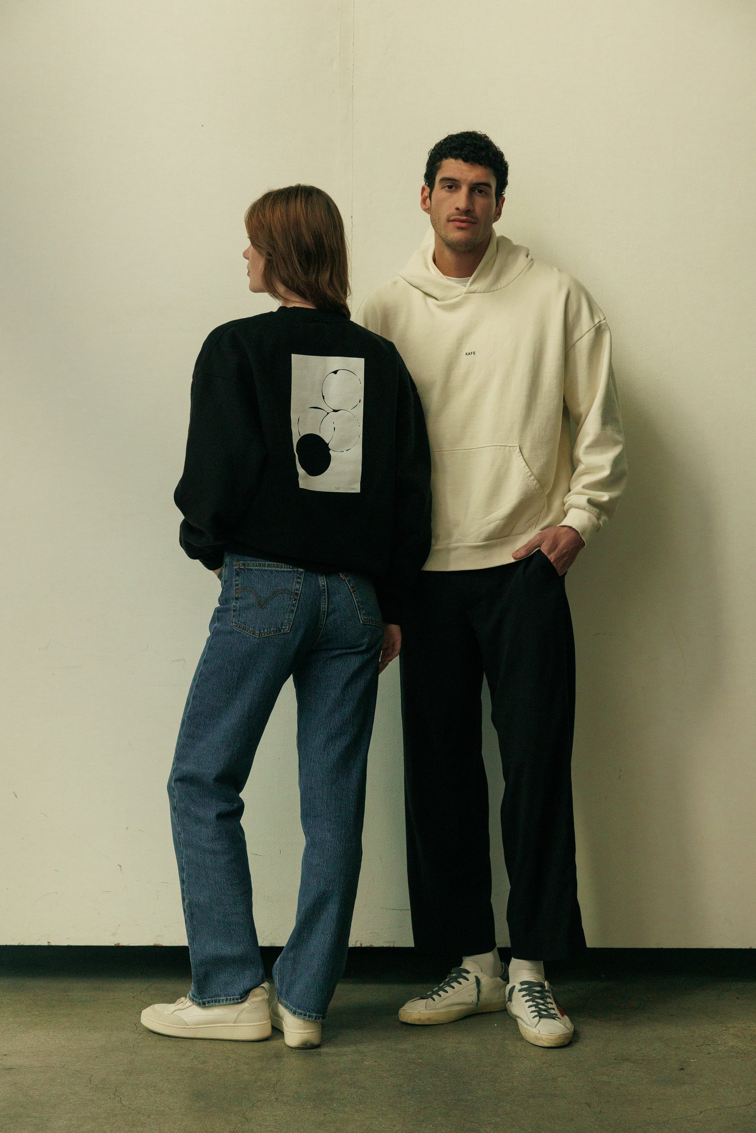 Two people standing against a plain wall, one wearing a black KAFE sweatshirt and, the other in a white KAFE hoodie.